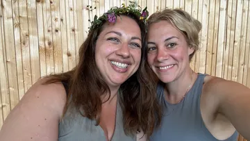 Zwei Frauen lächeln, sitzen vor Holzwand.