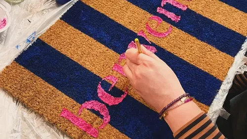 Hand paints doormat with pink text.