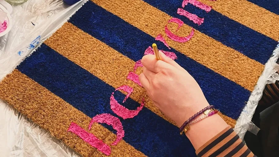 Hand paints doormat with pink text.