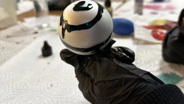 Behandschuhte Hand hält bemalten Ball drinnen.