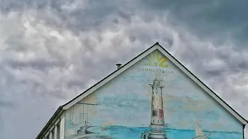 Gebäudewand mit Leuchtturm-Wandbild, wolkiger Himmel.
