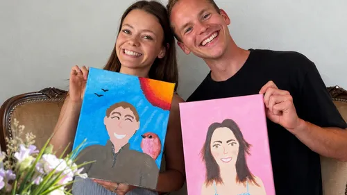 Two people smiling, holding painted portraits indoors.