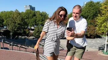 Zwei Personen schauen auf Handy, Park im Hintergrund.