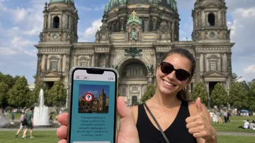 Frau lächelt, hält Telefon, Berliner Dom im Hintergrund.