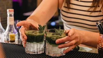 Hands holding two green drinks on bar.