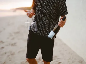 Man carries bottle and glasses on the beach.