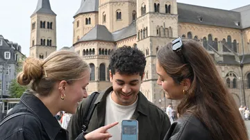 Drei Personen betrachten Handy vor historischer Kulisse.