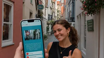 Person hält Handy, lächelt in schmaler Straße.