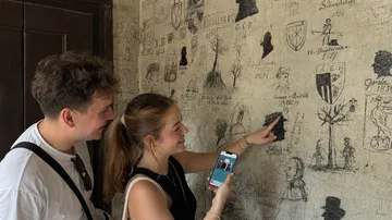Zwei Personen betrachten Wand mit historischem Graffiti.