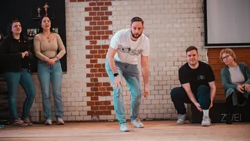 Person crouching, five others watch, brick wall backdrop.