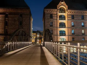 Holzbrücke verbindet zwei Backsteingebäude bei Nacht.