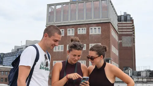 Drei Personen schauen auf Handy außerhalb des Gebäudes.