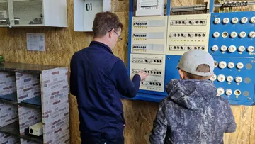 Two people examine electric control panel.