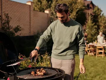 A man is grilling chicken in the garden, surrounded by guests.