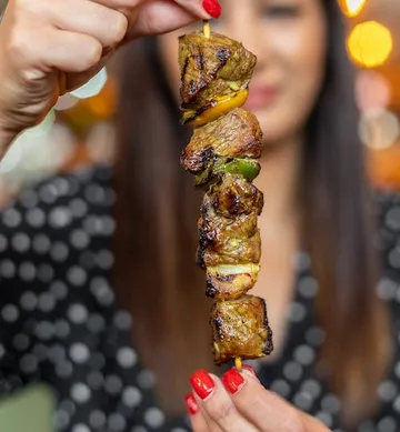 Gegrillter Fleischspieß, gehalten von einer Frau, Hintergündung unscharf.