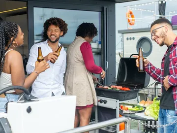 Grillende Freunde genießen Getränke auf einer Terrasse am Wasser.