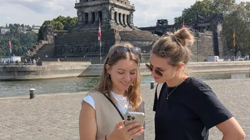 Zwei Frauen sehen aufs Handy vor Denkmal.
