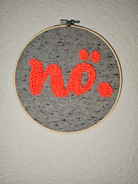 Stickrahmen mit rotem Text 'nö.' an Wand.