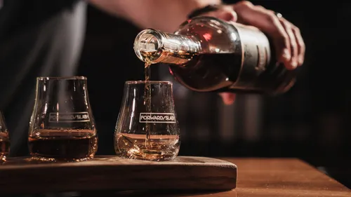 Hand gießt Whiskey in ein Glas auf einem Tisch.