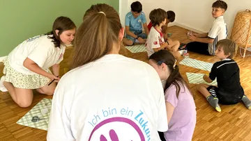 Kinder sitzen auf Boden, Erwachsene leiten.