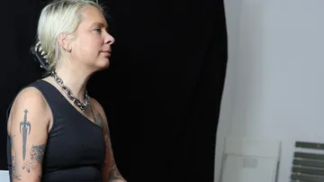 Person sitting, wearing a necklace, against black backdrop.