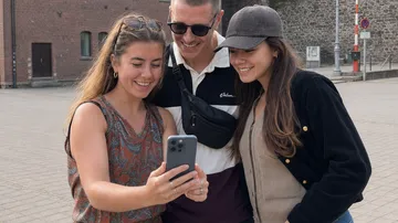Three people smiling at a smartphone outdoors.