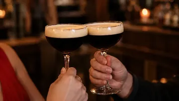 Zwei Cocktailgläser stoßen an; Barhintergrund.