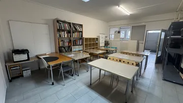 Klassenzimmer mit Schreibtischen, Stühlen, Regalen, und Ausrüstung.