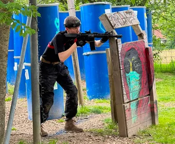Person zielt mit Paintball-Gewehr im Freien, Fässer im Hintergrund.