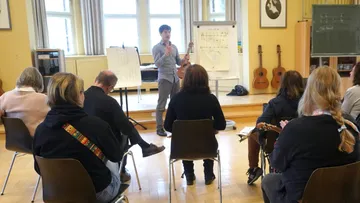 Person unterrichtet Gitarrenkurs mit sitzenden Schülern.