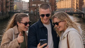 Three friends smiling at phone near river.