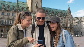 Drei Personen lächeln und schauen auf ein Handy vor einem historischen Gebäude.