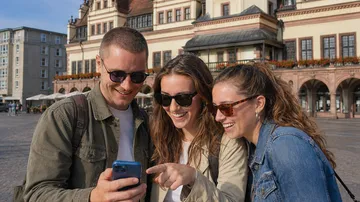 Three friends smiling, looking at phone outdoors.