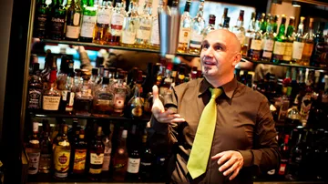 Bartender tossing shaker in liquor-filled bar.