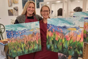 Two women holding colorful landscape paintings.