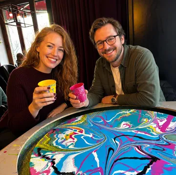 Zwei Personen halten Tassen neben bunter Kunst.