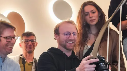 Four people look at camera screen indoors.