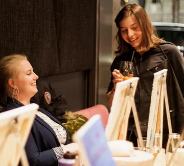 Zwei Frauen lachen, halten Getränke drinnen.