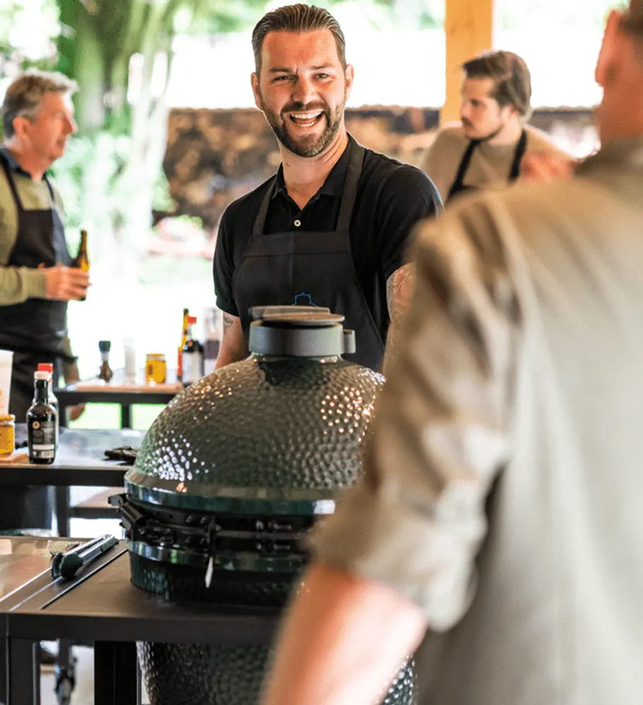 Man smiles while barbecuing, surrounded by friends and nature.