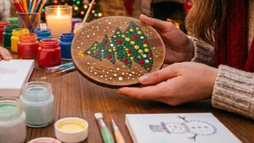 Hands holding painted Christmas tree plate, surrounded by paints.