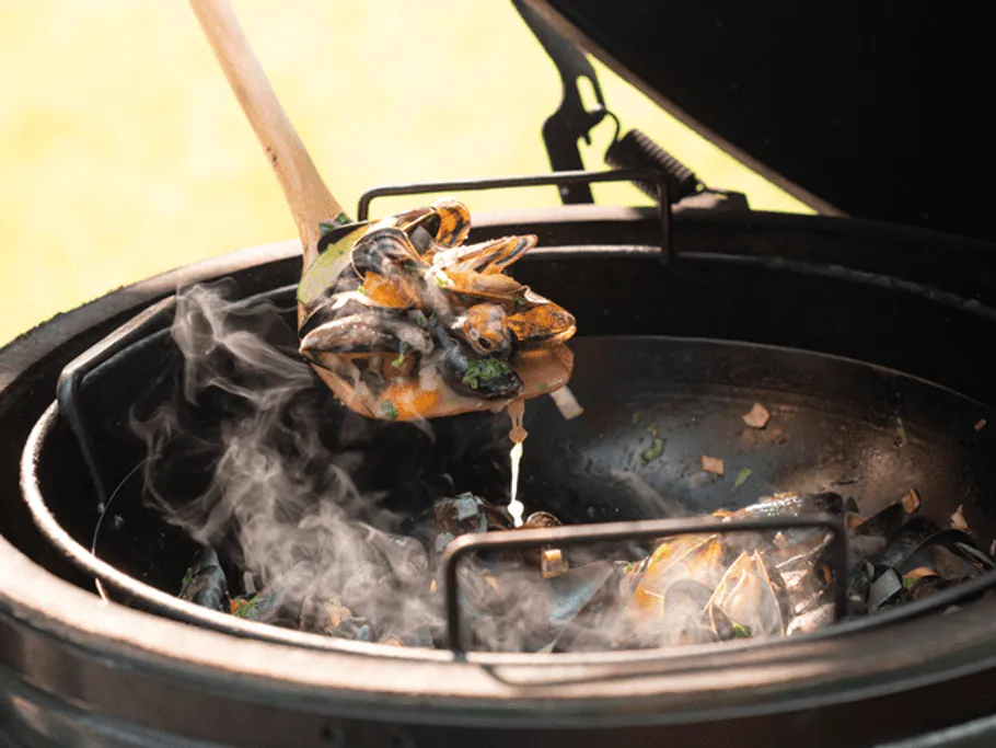 Mussels are lifted out of a smoking grill with a spoon.