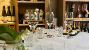 Wine glasses and cheese on table, shelves with bottles.