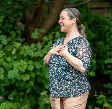 Woman smiling, hands on chest, green outdoors.