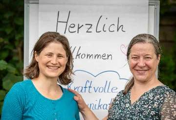 Two women smiling beside welcome sign outdoors.