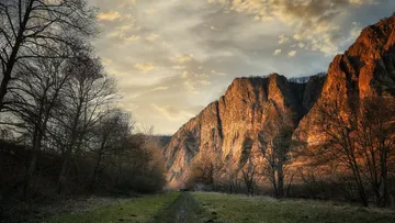 Sunlit rocky mountains, trees, grassy path.