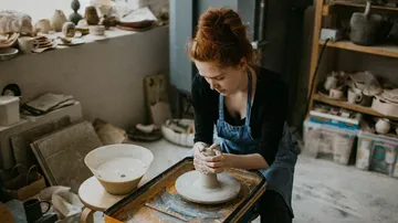Person shaping clay on a pottery wheel inside studio.