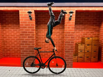 Person handstands on bicycle in a red-brick room.