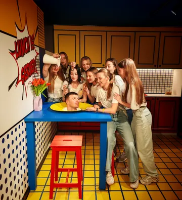 Männerkopf auf Tisch, Frauen drumherum, spielerische Szene.