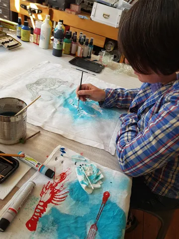 Person painting on fabric, surrounded by paints.