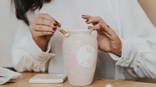 Person painting vase at wooden table.